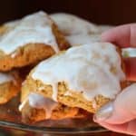 Hand holding an iced pumpkin scone. There is a pile of pumpkin scones in the background.