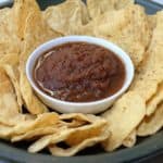 A small white bowl of dark red salsa surrounded by corn tortilla chips.