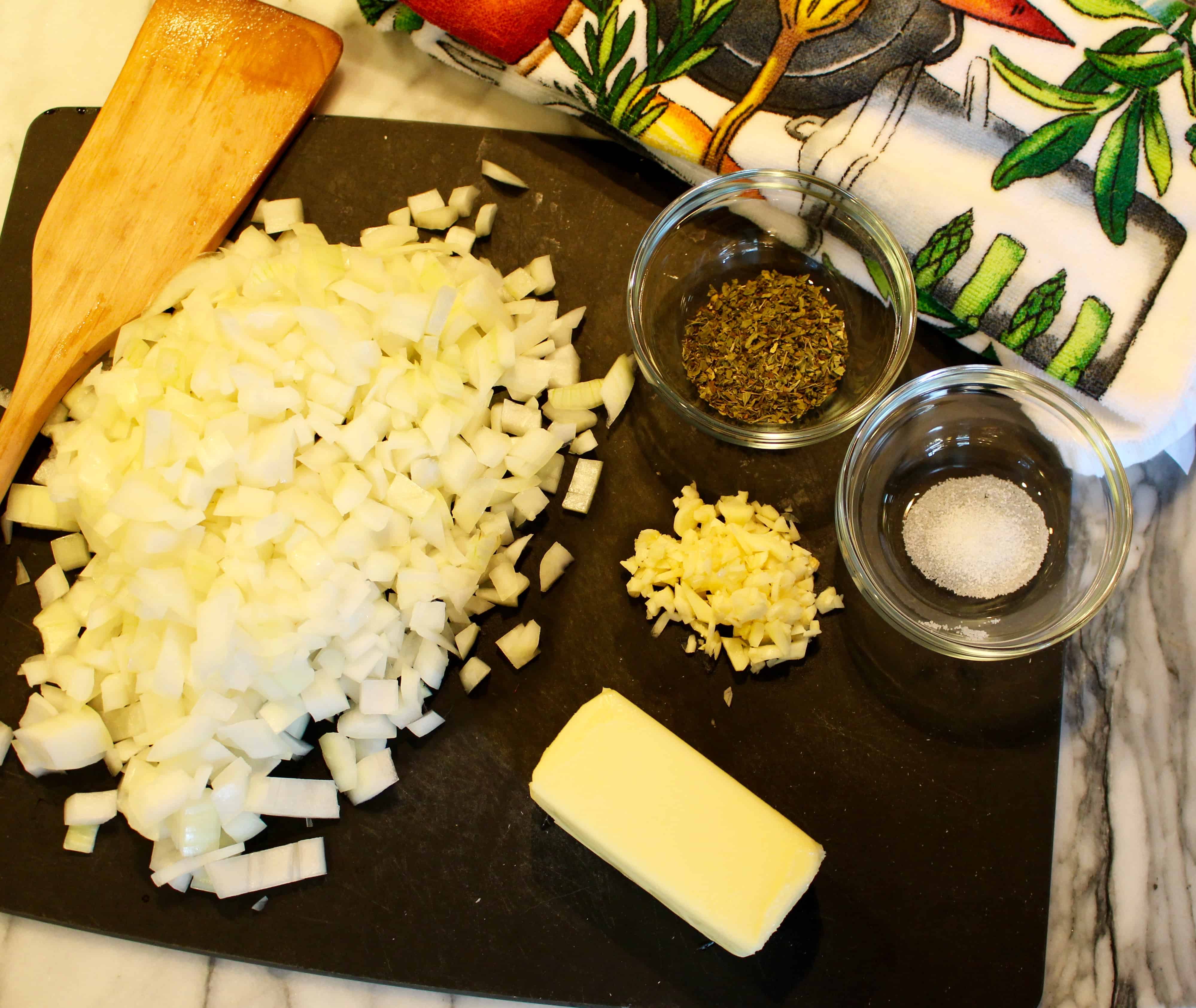 Chopped onion, chopped garlic, a small bowl with salt, a small bowl with oregano, a cube of butter and a wooden spatula on a black cutting board on a marble background next to a kitchen towel with leaves on it.