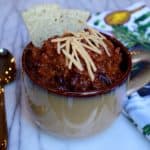 Large cup of chili with cheddar cheese on top and 2 corn chips stuck in the back of the cup. A spoon is on the left next to the cup of chili and a kitchen towel is on the right.