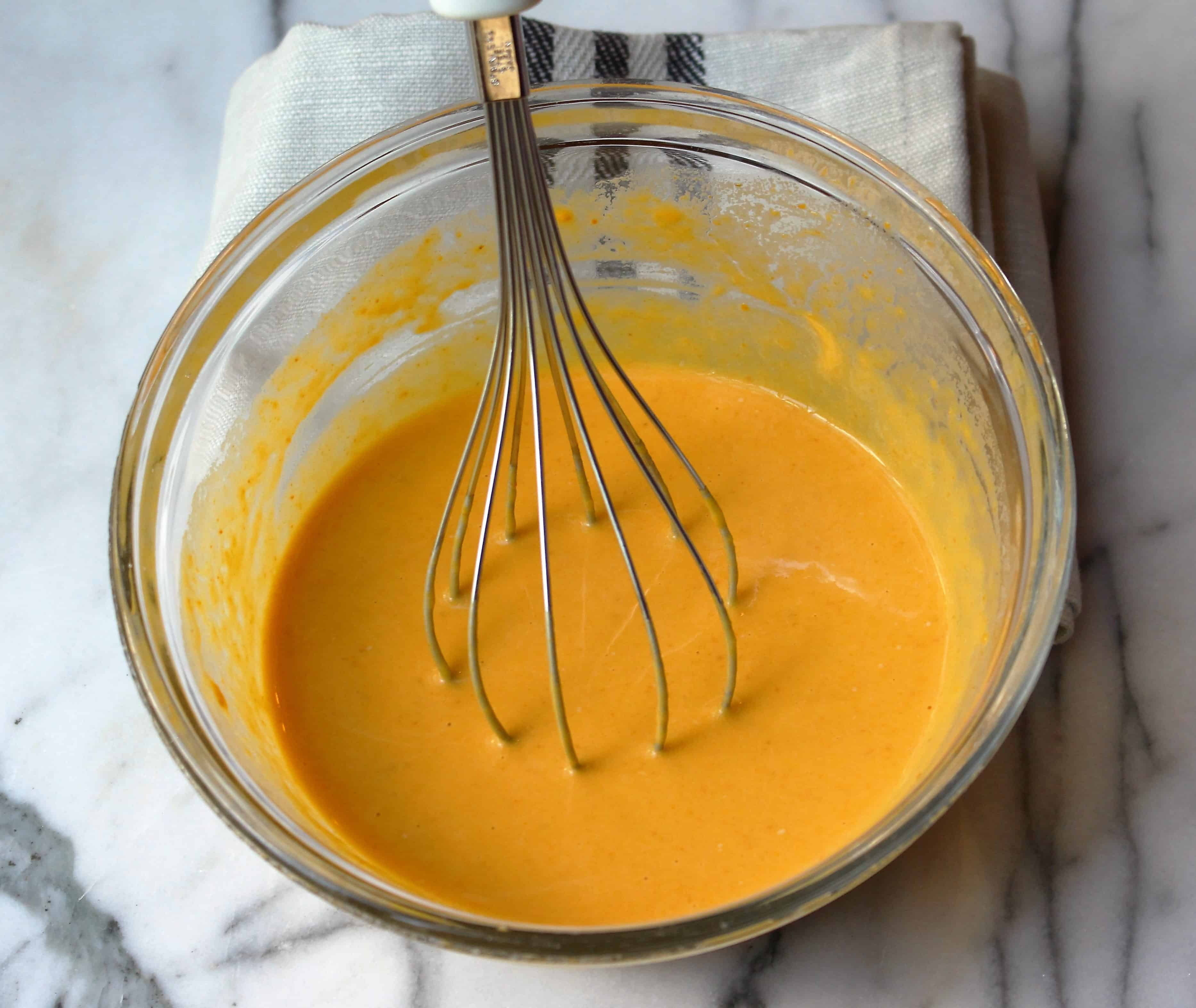 Whisk, pumpkin puree, half and half and egg mixture in a glass bowl on a marble background. A grey kitchen towel with black stripes behind the bowl.