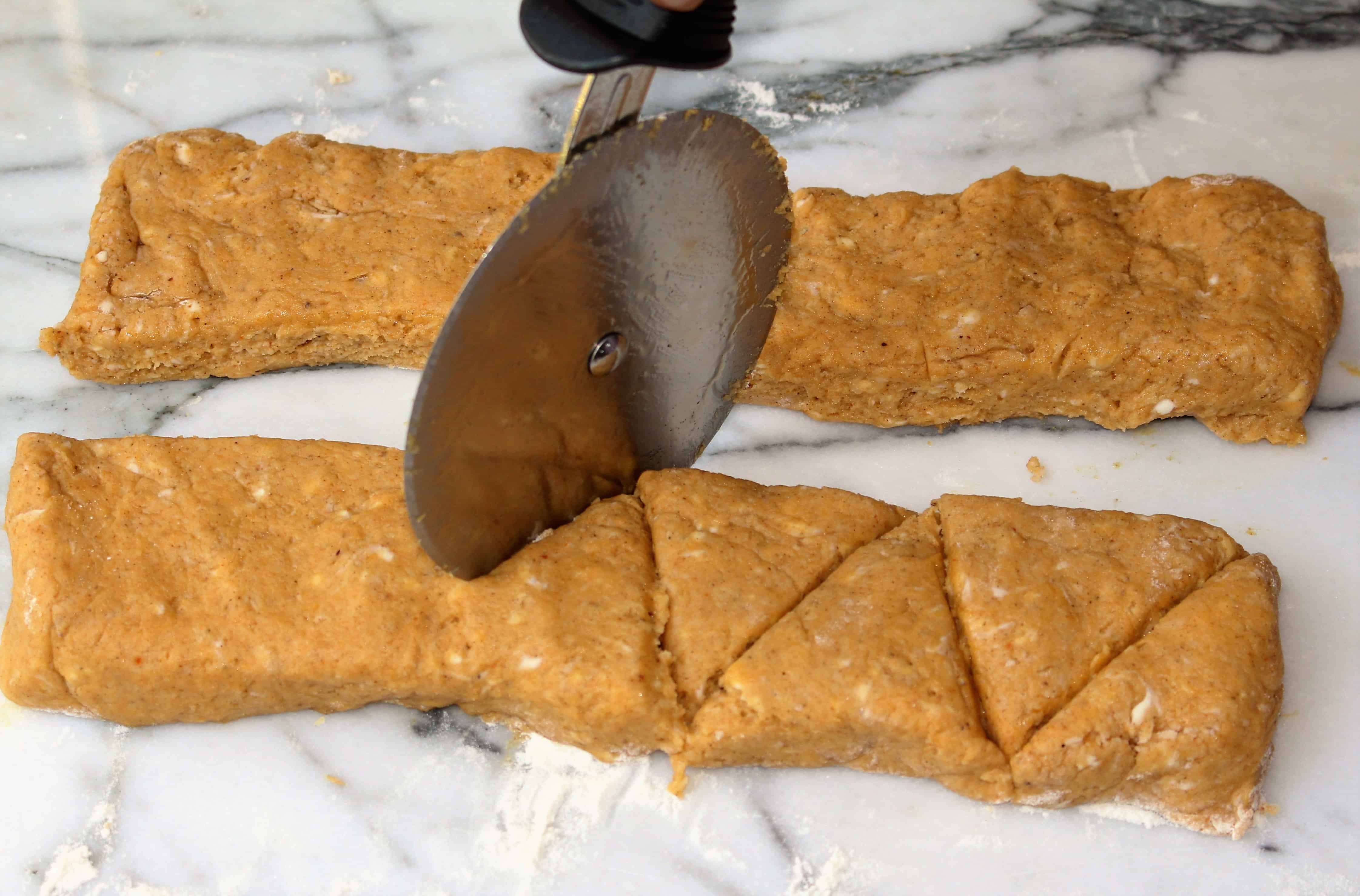 Two pieces of pumpkin scone dough formed into rectangles lying on a marble background. A pizza slices is slicing through one of the rectangles creating triangles.