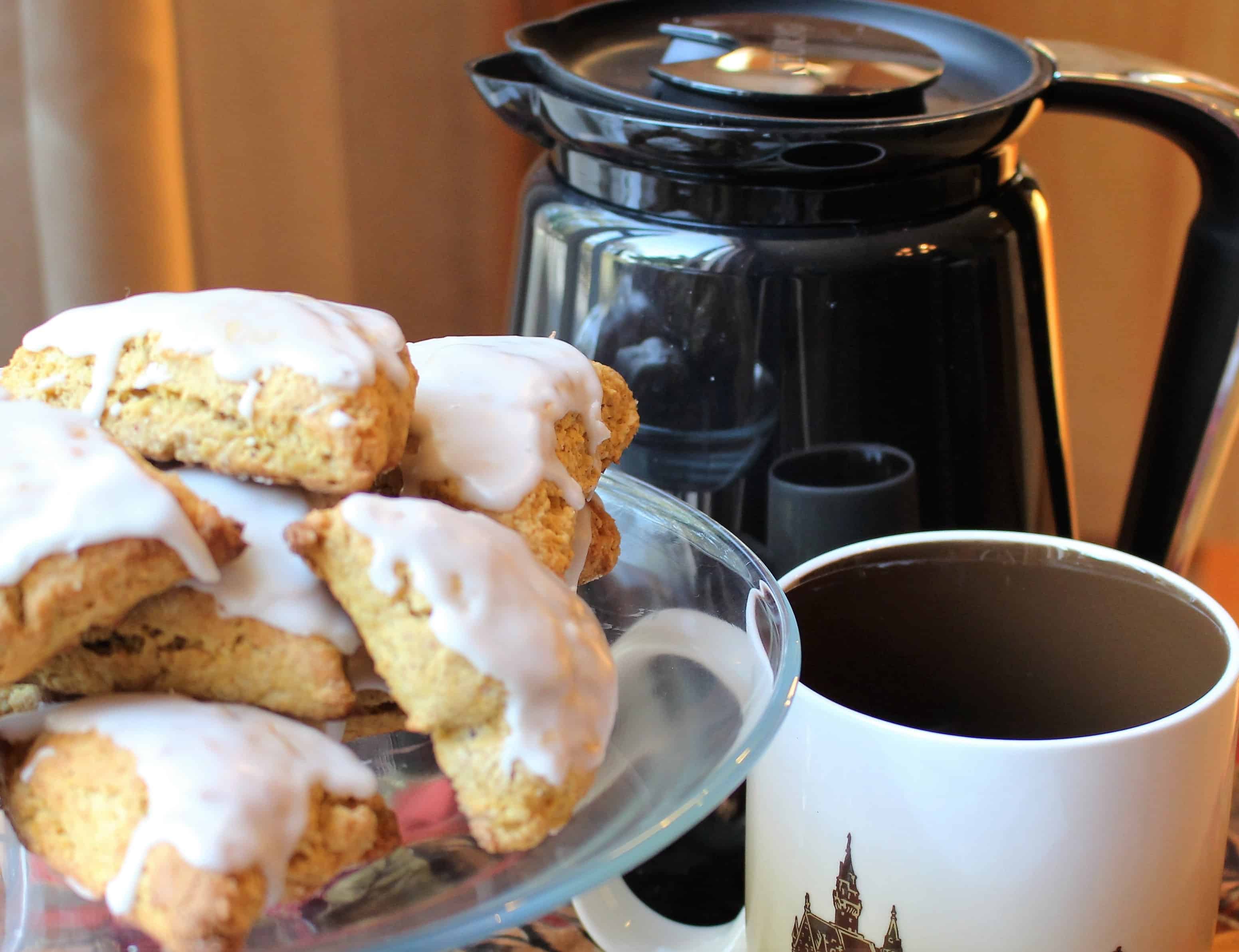 A glass plate with a pile of iced pumpkin scones on it. A white and brown coffee mug next to the plate. A black coffee pot is behind the plate and coffee mug.