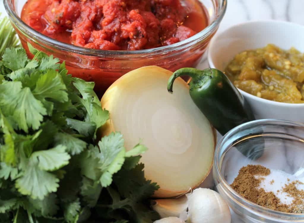 A bunch of cilantro, half a yellow onion, garlic, a small glass bowl of spices, green jalapeno pepper, small white bowl of diced green chilis and a medium glass bowl of fire roasted tomatoes on a marble background.