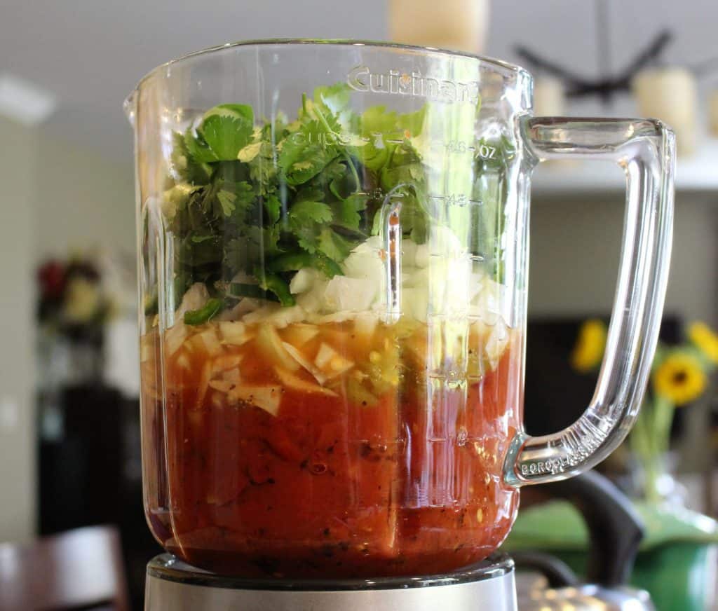 A bunch of cilantro, chopped onion, garlic, spices, green jalapeno pepper,  diced green chilis, chipotle peppers and fire roasted tomatoes in a glass blender.