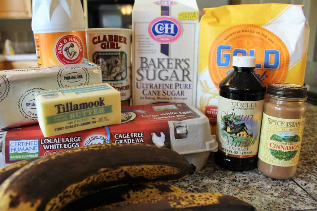 Brown bananas, egg carton, cube of butter, cube of cream cheese, vanilla extract, cinnamon, flour, sugar, baking powder and baking soda lying on a speckled counter top.