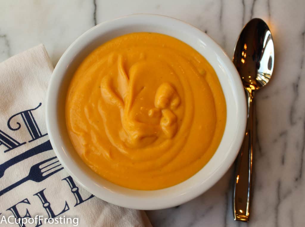 Butternut Squash Soup in a white bowl with a silver spoon on the right and a kitchen towel on the left. This is all on top of a marble background.
