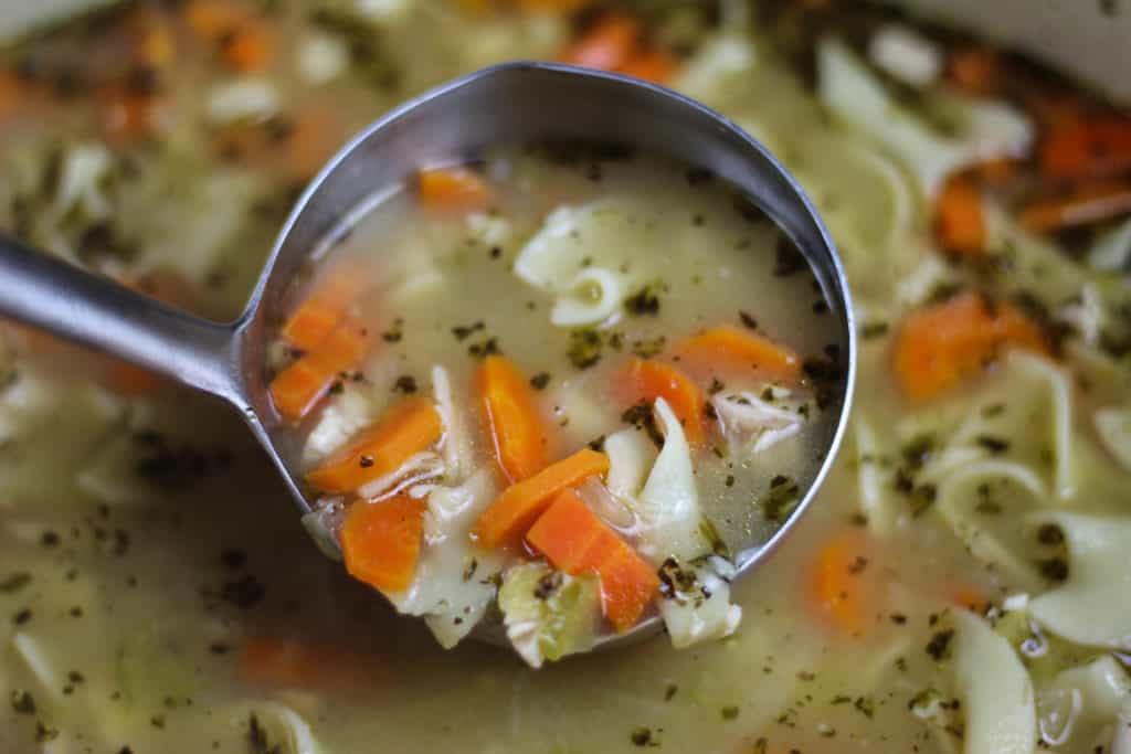 A ladle full of chicken soup being scooped from the crock pot full of soup. You can see the noodles, celery, carrot slices, chicken and fresh spices.