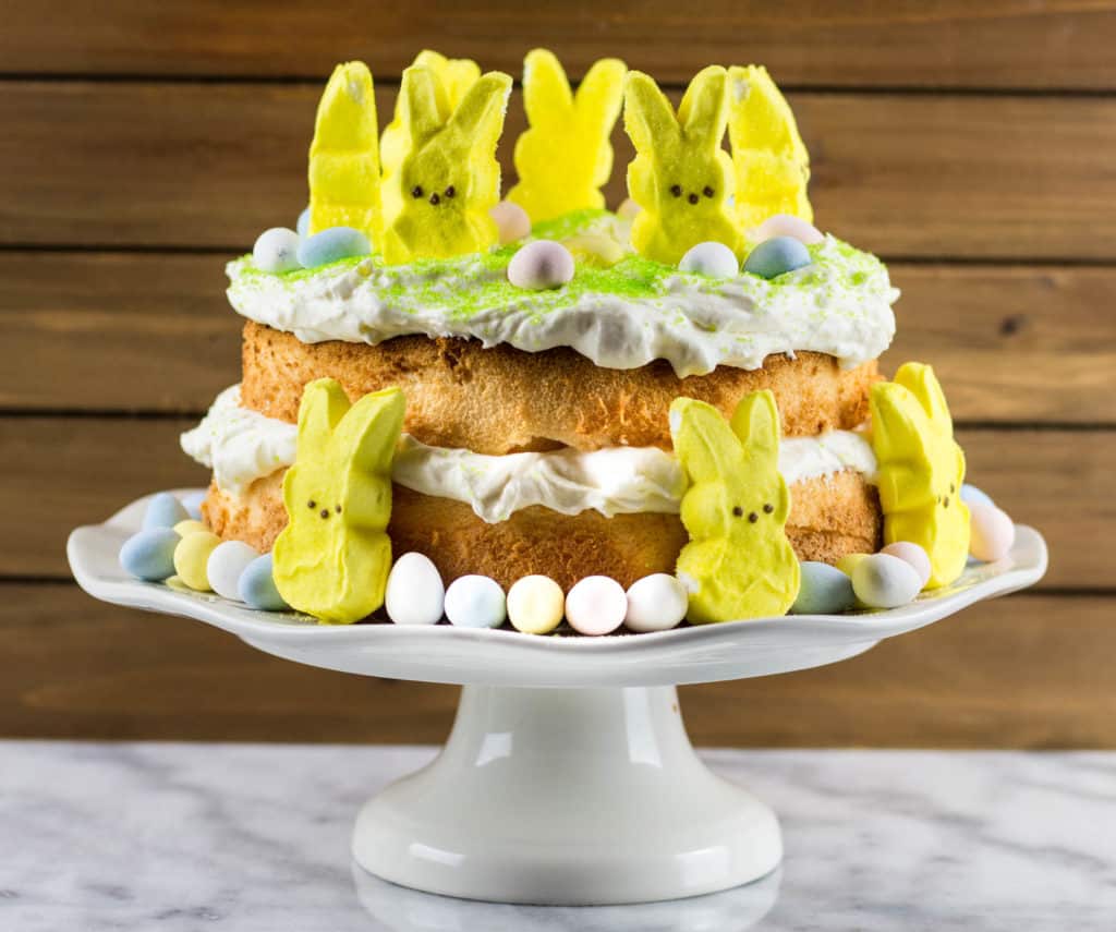 Angel food cake cut in half  horizontally with lemon curd filling in the middle of the halves and on top of the cake. There are yellow peeps on top and around the sides of the cake. Also, there are candied eggs on top and around the cake and green sprinkles.