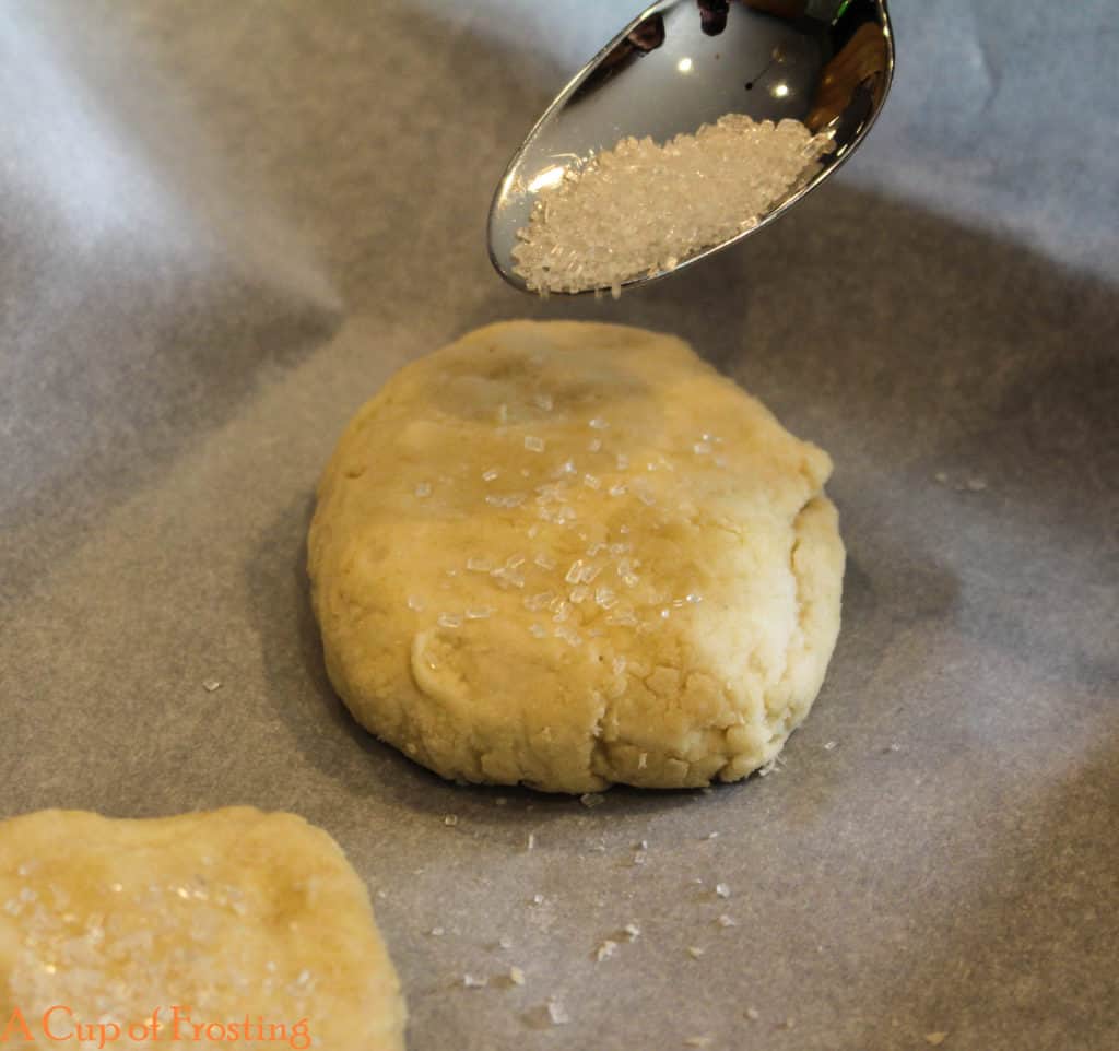 Sprinkling course sugar on a flattened small round ball of shortcake biscuit dough sitting next to another sugared biscuit on top of parchment paper.