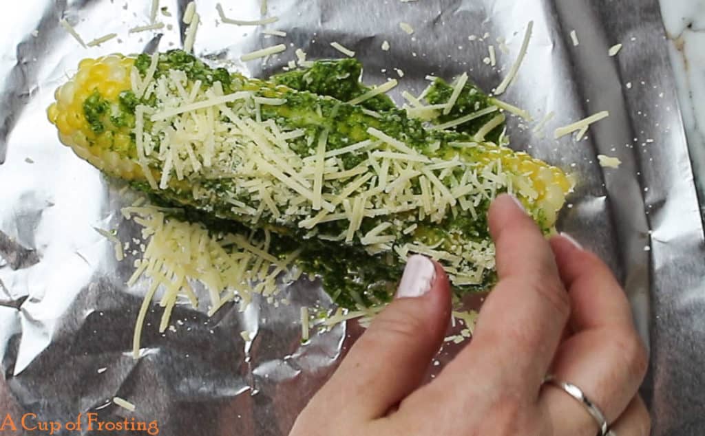 Corn on the cob with green pesto and shredded parmesan on top lying on a piece of foil.