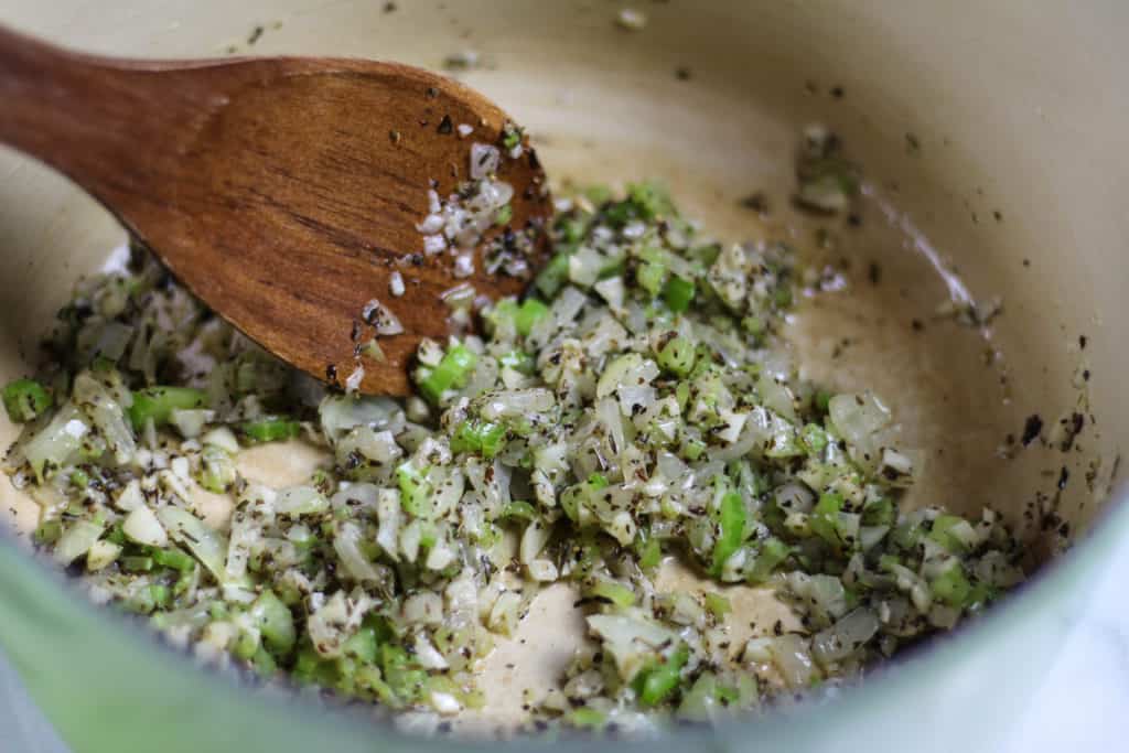 Minced garlic, chopped onion and chopped celery with salt and pepper in a large crock pot pan being stirred with a wooden spoon.