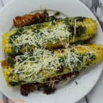 Corn on the cob with green pesto and shredded parmesan on top lying on a white dinner plate. There are crispy looking pieces of shredded parmesan under and beside the corn on the cob.