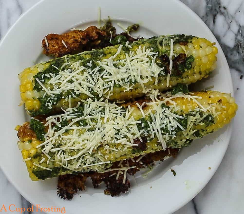 Corn on the cob with green pesto and shredded parmesan on top lying on a white dinner plate. There are crispy looking pieces of shredded parmesan under and beside the corn on the cob.