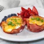 Two cooked ham and egg cups with melted cheese and slices of green onion on top. This is on a white medium sized plate with blueberries and sliced strawberries. There is a grey dishtowel with 3 stripes on the bottom with a silver fork on top to the left of the white plate. The background is white marble. - 1
