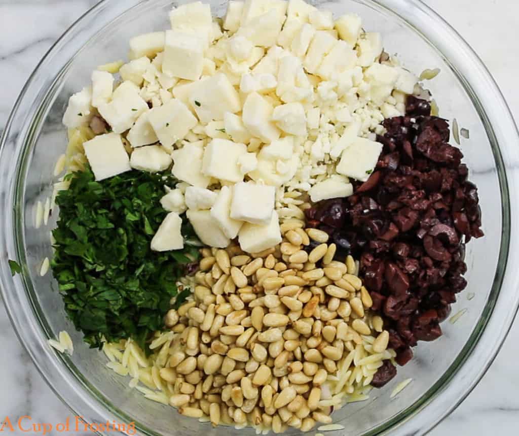 A medium glass bowl filled with cooked orzo topped with feta cheese crumbles, chopped Kalamata olives, chopped Italian parsley and toasted pinenuts.