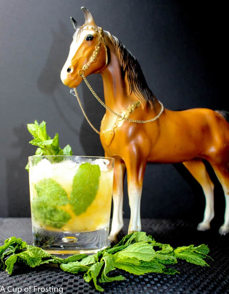 Mint Julep in a cocktail glass with a sprig of mint in the top of the glass and mint leaves showing inside the liquid in the glass. A toy hors that is golden brown is standing next to the glass and there is a sprig of mint in front of the glass.