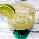 A lime green margarita with ice cubes in a margarita glass with a lime wedge on the side of the glass.
