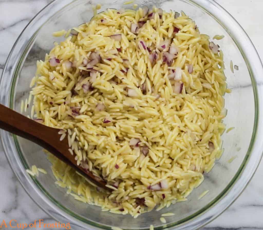 A medium glass bowl filled with cooked orzo with finely chopped red onions being stirred by a wooden spoon on a white marble background.