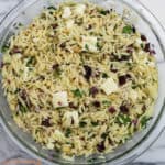 A medium glass bowl filled with cooked orzo with feta cheese crumbles, chopped Kalamata olives, chopped Italian parsley and toasted pinenuts.
