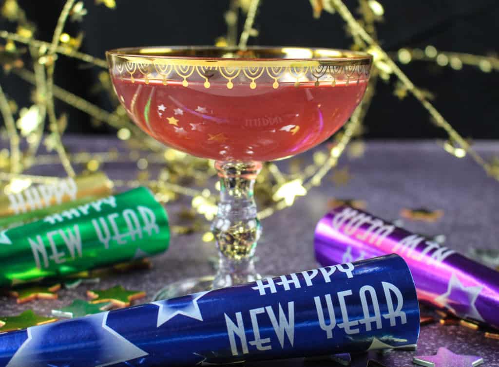 A decorative gold rimmed wine glass with Pomegranate, Lime and Champagne Cocktail. There are New Year's horns in different colors lying around the cup and gold decorations in the background.