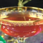 A decorative gold rimmed wine glass with Pomegranate, Lime and Champagne Cocktail. There are New Year's horns in different colors lying around the cup and gold decorations in the background.