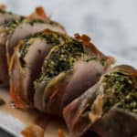 Side shot of a cooked and sliced pork tenderloin wrapped with the herb mixture and prosciutto sitting on a white oblong serving plate on a white marble background.