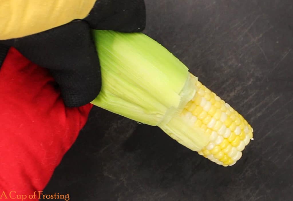 A yellow and black oven mitt is holding a corn on the cob, which is being shucked. There is a red towel lying under the corn on the cob.