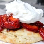 Sliced strawberries sitting on top of a cooked biscuit with balsamic vinegar dripping down the sides and whipped topping on top of the strawberries.